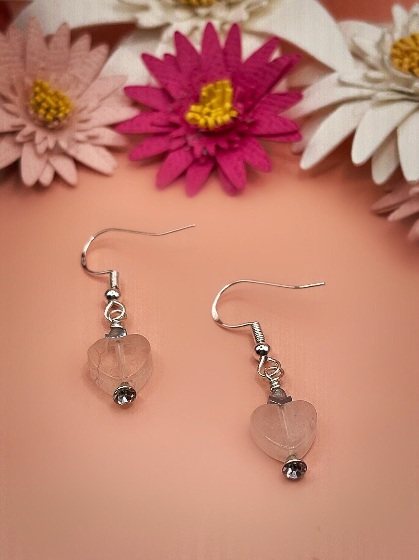 Peach background with paper daisies in hot pink and baby pink and white. There are two silver earring hooks with one inch earring drop with a rose Quartz heart bead, small silver spacer bead, and small labradorite bead on top.