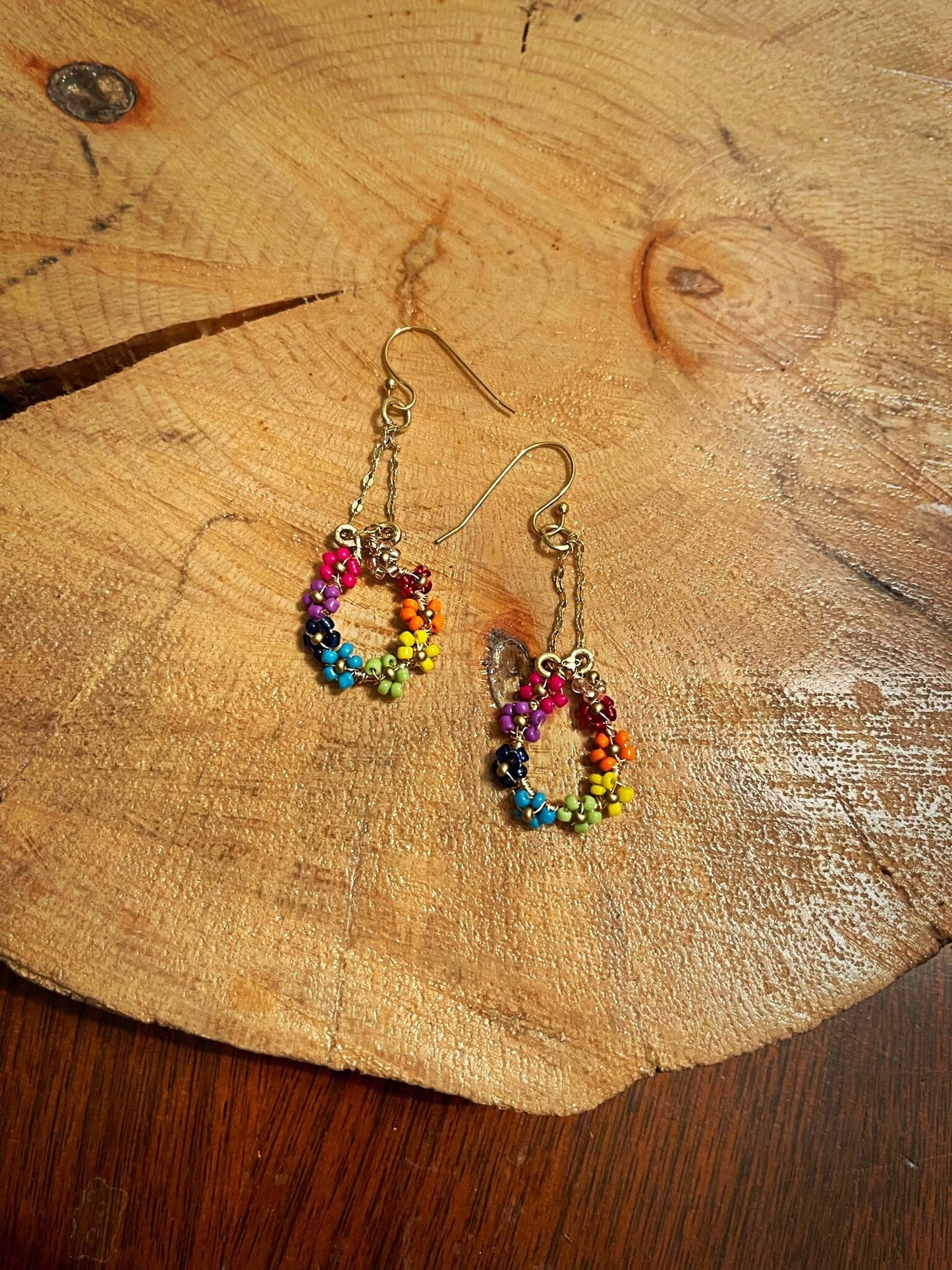 Beaded flower rainbow earrings with bright colors and small seed bead flowers circling around an oval of gold wire hanging from gold chain on a gold earring hook